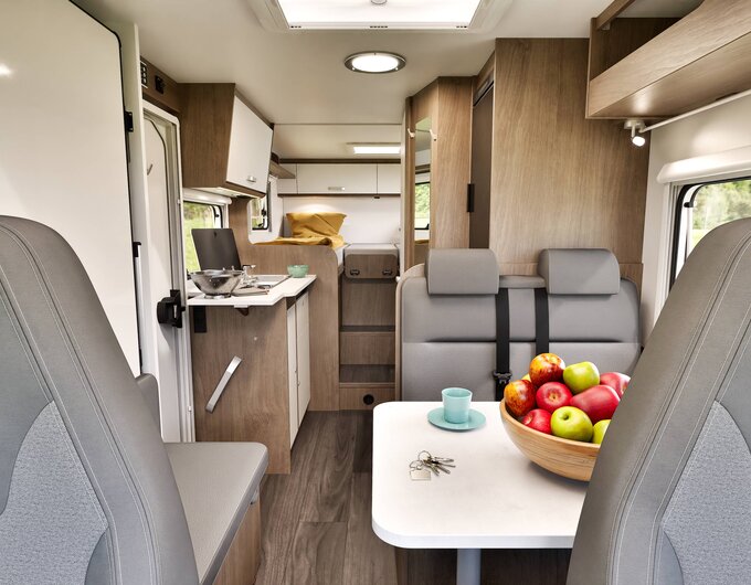 Carado motorhome interior with a seating area, table, fruit bowl, compact kitchen and rear bed
