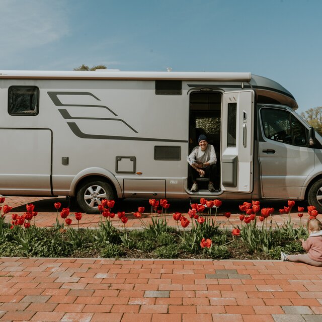 Side view of a HYMER motorhome with a young man sitting in the open cabin door; red tulips in the foreground