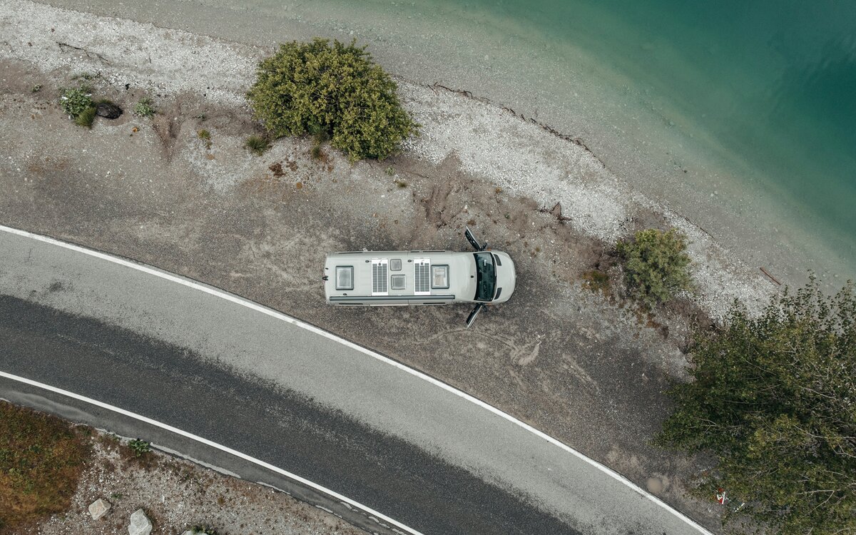 Fourgon gris vu d'en haut, stationné sur du gravier à côté d'un lac au bord de l'eau, à côté du gravier, il y a une petite route et sur le lit de gravier, il y a trois arbres verts, les portes du fourgon sont ouvertes