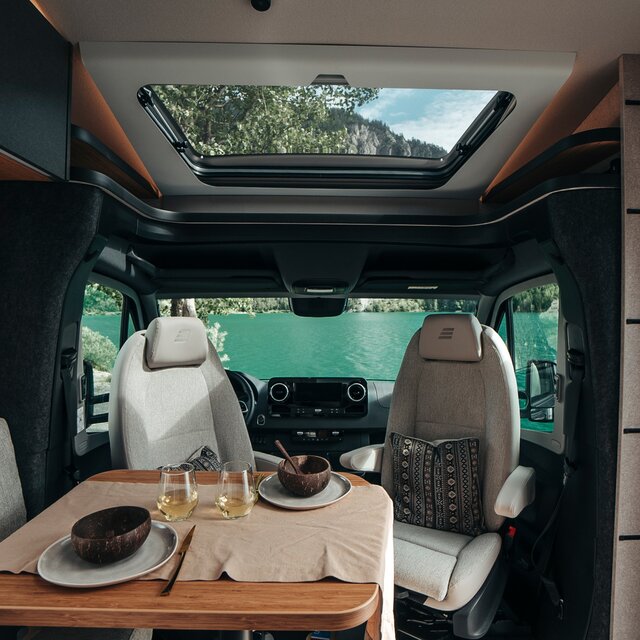 Bordet i HYMER ML-T i stildesignen Native Bamboo är dukat med tallrikar och vitt vin. Bakom finns förarhytten med utsikt över naturen.
