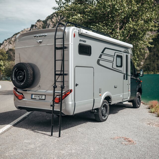 HYMER ML-T syns från en sned vinkel bakifrån. Den står lätt vriden framför en bergssjö och takstegen är utfälld.