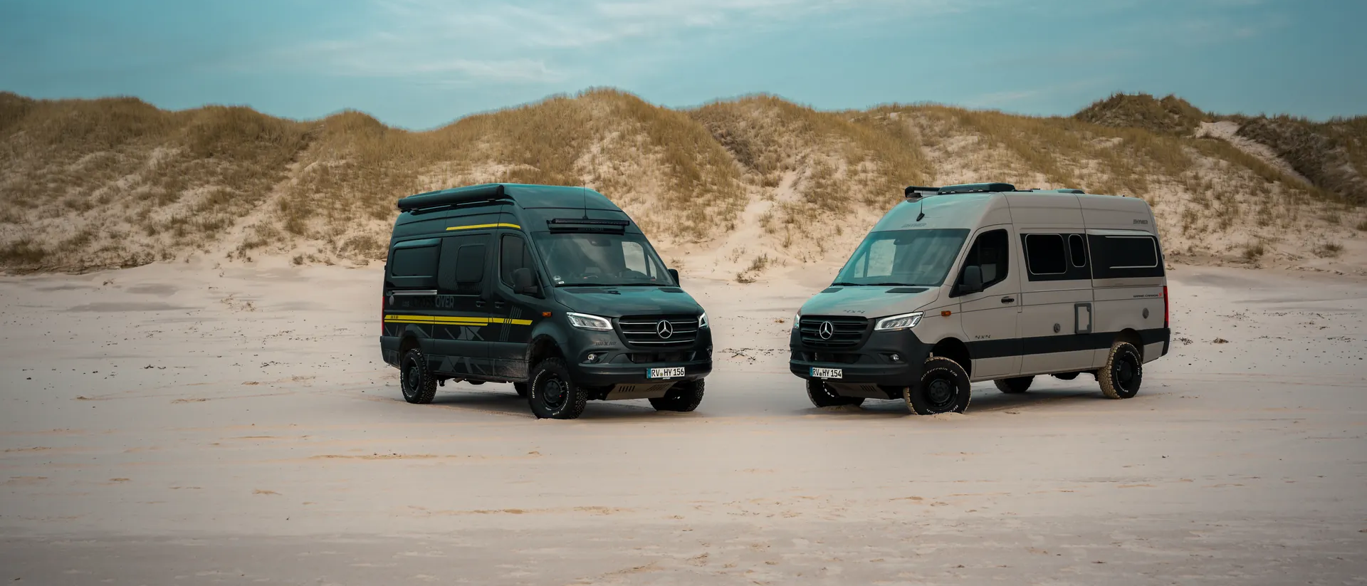 A black and a stone-gray campervan on a Mercedes chassis at a sandy beach with dunes in the background	
